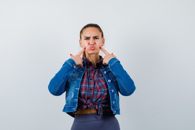 Jeune femme pointant du doigt les joues gonflées, faisant la moue des lèvres en chemise, veste et l'air déçue, vue de face.