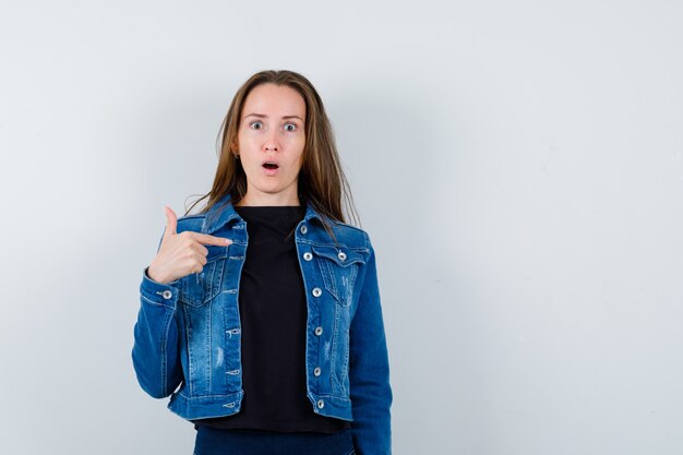 Jeune femme pointant sur elle-même en blouse, veste et à la perplexité, vue de face.