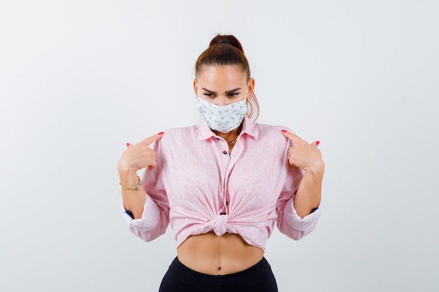 Jeune femme pointant sur elle-même en chemise, pantalon, masque médical et à la vue de face, hésitante.