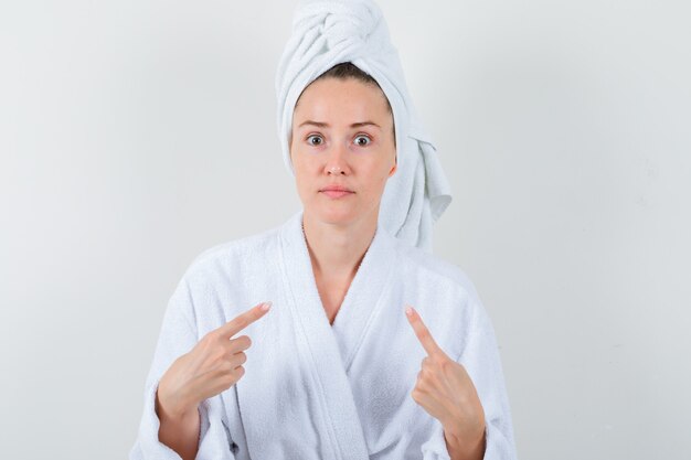 Jeune femme pointant sur elle-même en peignoir blanc, serviette et à la perplexité, vue de face.