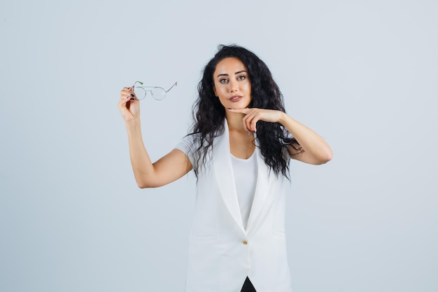 Photo gratuite jeune femme pointant sur des lunettes en t-shirt blanc, veste et l'air confiant, vue de face.