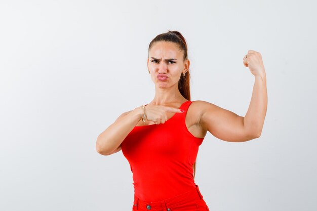 Jeune femme pointant sur les muscles du bras en débardeur rouge, pantalon et à la vue de face, confiant.