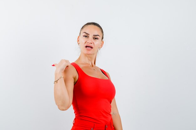 Jeune femme pointant le pouce vers le côté gauche en débardeur rouge, pantalon et à la confiance. vue de face.