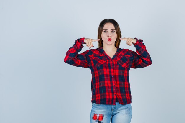 Jeune femme pointant sur sa tête, faisant la moue en chemise à carreaux, jeans et semblant positive. vue de face.