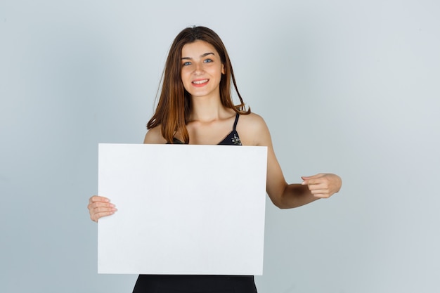 Jeune femme pointant sur une toile vierge en chemisier et semblant joyeuse. vue de face.