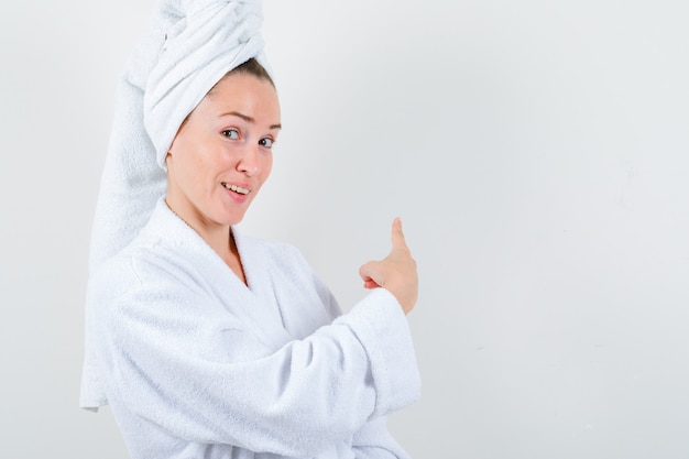 Jeune femme pointant vers l'arrière en peignoir blanc, serviette et à la joyeuse. vue de face.