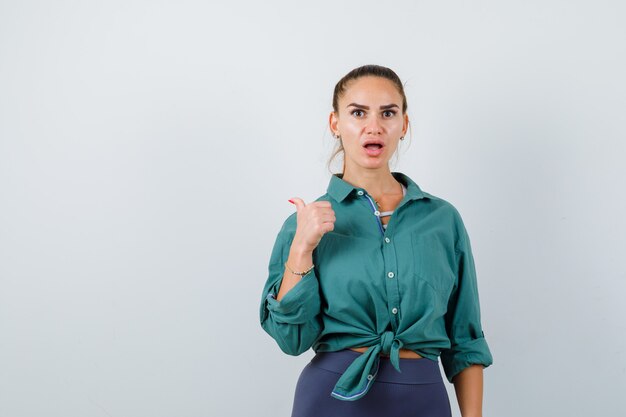 Jeune femme pointant vers l'arrière avec le pouce en chemise verte et l'air surpris, vue de face.