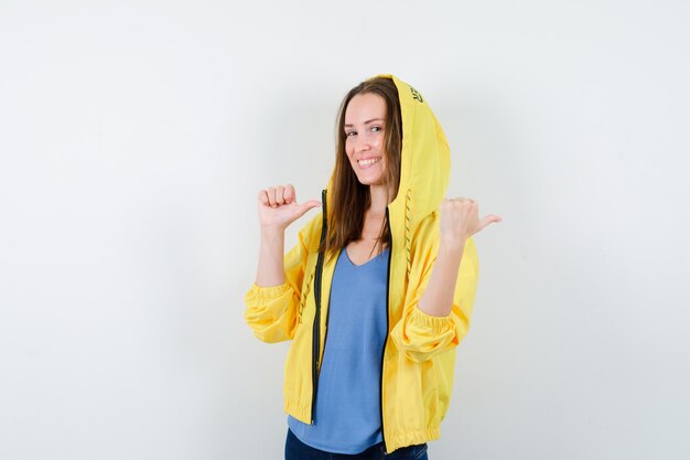 Jeune femme pointant vers l'arrière avec les pouces et l'air joyeux, vue de face.