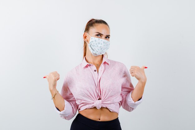 Jeune femme pointant vers l'arrière avec les pouces en chemise, masque et à la vue confiante, de face.