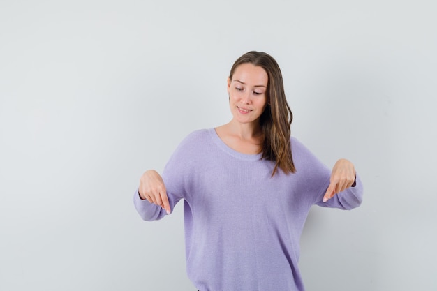 Jeune femme pointant vers le bas en blouse lilas et à la recherche concentrée. vue de face.