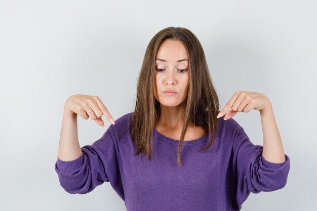 Jeune femme pointant vers le bas en chemise violette et à la recherche concentrée. vue de face.