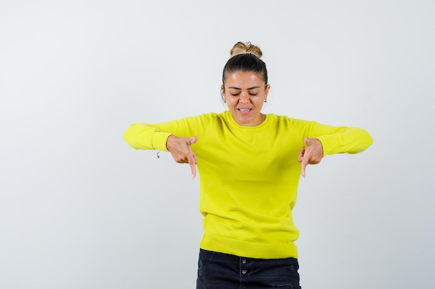 Jeune femme pointant vers le bas avec l'index en pull jaune et pantalon noir et à heureux