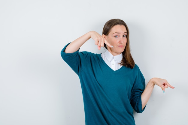 Photo gratuite jeune femme pointant vers le bas en pull sur chemise blanche et à la vue de face indécise.