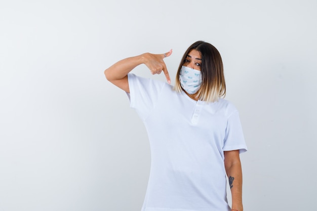 Jeune femme pointant vers le bas en t-shirt, masque et à la vue de face, confiant.