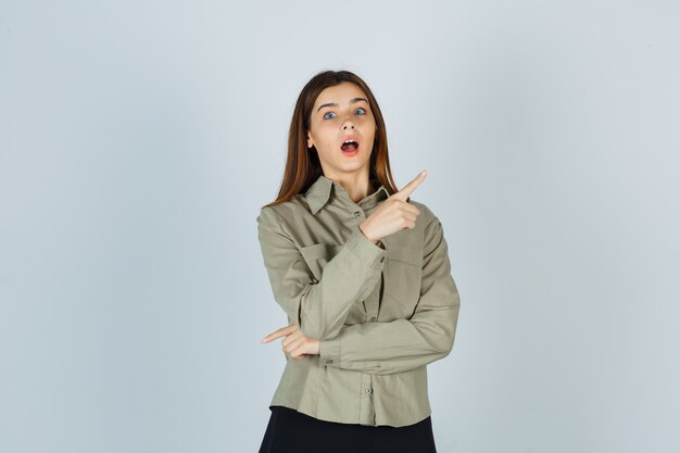 Jeune femme pointant vers le coin supérieur droit en chemise, jupe et se demandant , vue de face.