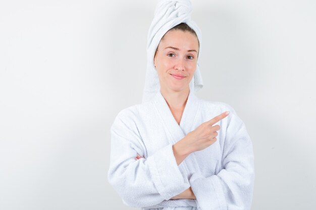 Jeune femme pointant vers le coin supérieur droit en peignoir blanc, serviette et à la satisfaction. vue de face.