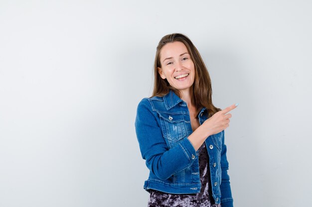 Jeune femme pointant vers le coin supérieur droit en veste en jean et l'air joyeux. vue de face.