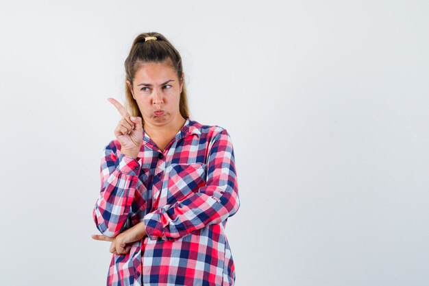 Jeune femme pointant vers le coin supérieur gauche en chemise à carreaux et à la vue de face, hésitante.