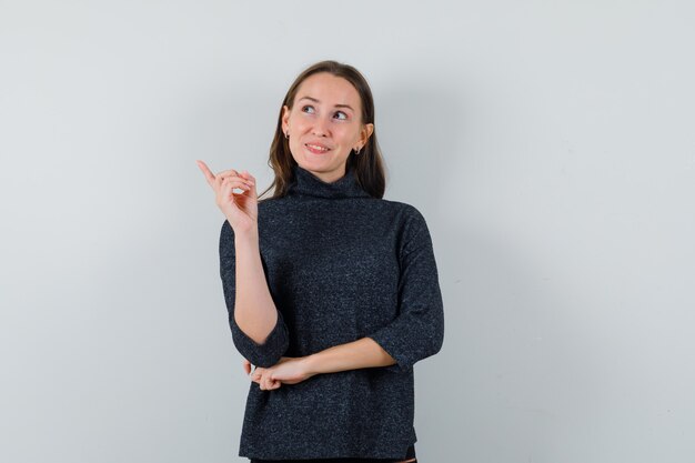 Jeune femme pointant vers le coin supérieur gauche en chemise décontractée et à la recherche d'espoir. vue de face.