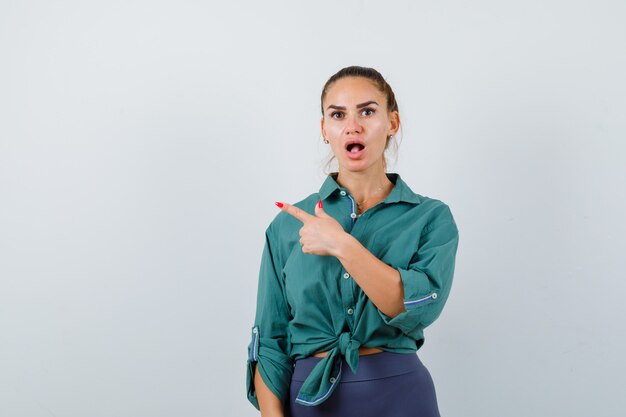 Jeune femme pointant vers le coin supérieur gauche en chemise verte et l'air choqué. vue de face.