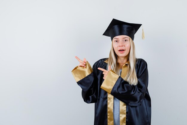 Jeune femme pointant vers le coin supérieur gauche en uniforme diplômé et semblant étonnée.