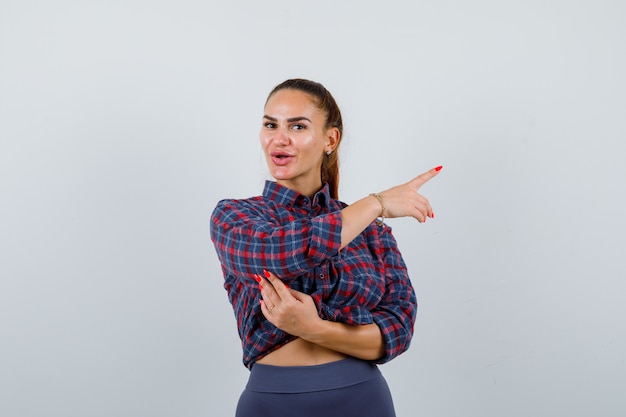 Jeune femme pointant vers le côté droit en chemise à carreaux, pantalon et à l'air confiant. vue de face.