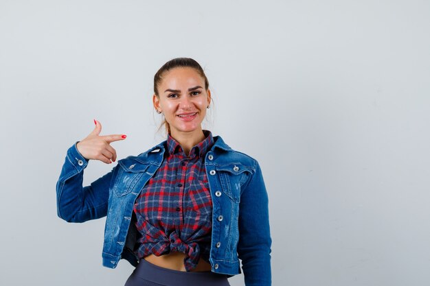 Jeune femme pointant vers le côté droit en chemise à carreaux, veste en jean et l'air joyeux. vue de face.