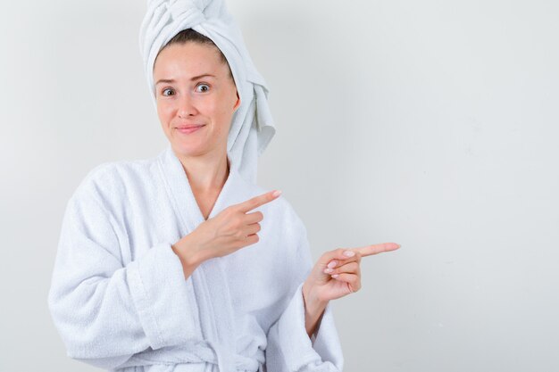 Jeune femme pointant vers le côté droit en peignoir blanc, serviette et à la drôle. vue de face.