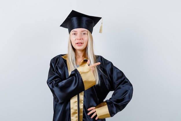 Jeune femme pointant vers le côté droit en tenue universitaire et semblant confiante.