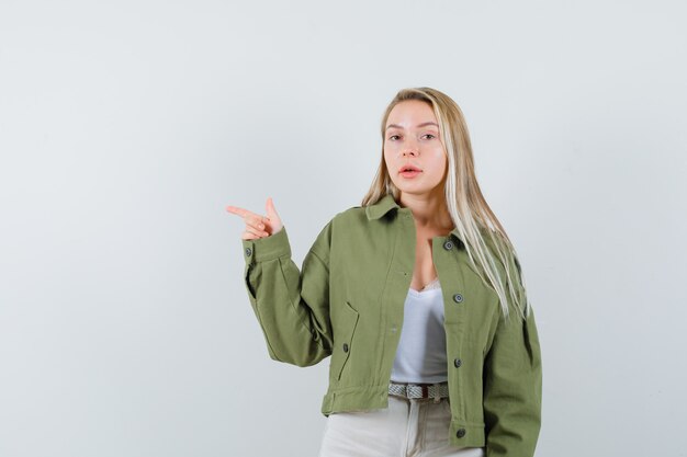 Jeune femme pointant vers le côté droit en veste, pantalon et regardant hésitante. vue de face.