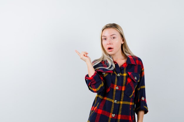 Jeune femme pointant vers le côté gauche en chemise à carreaux et regardant perplexe, vue de face.