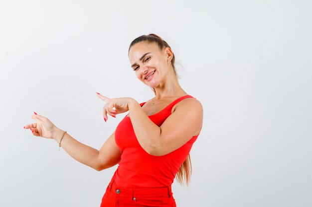 Jeune femme pointant vers le côté gauche en débardeur rouge, pantalon et air heureux. vue de face.