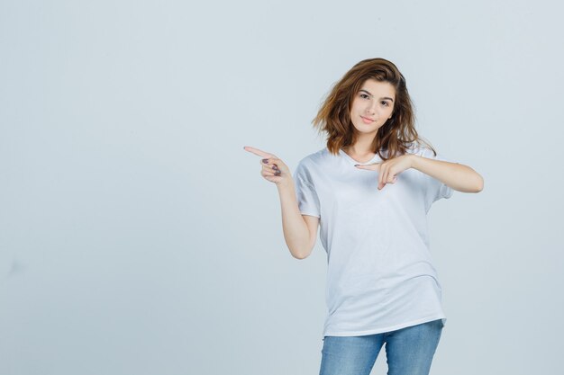 Jeune femme pointant vers le côté gauche en t-shirt, jeans et regardant confiant, vue de face.