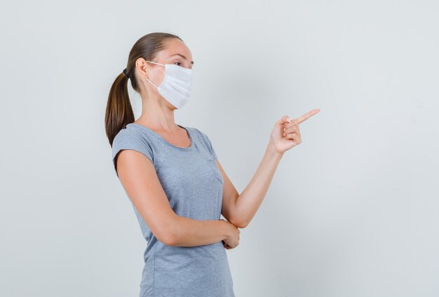 Jeune femme pointant vers le côté en t-shirt gris, masque et à la recherche concentrée. vue de face.