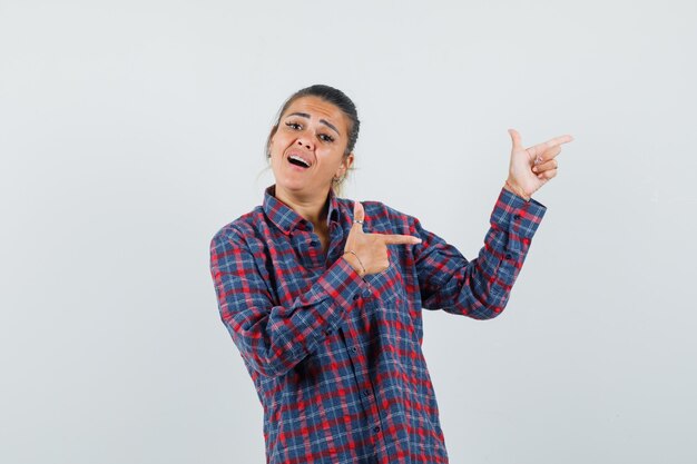 Jeune femme pointant vers la droite avec l'index en chemise à carreaux et à la pensif. vue de face.