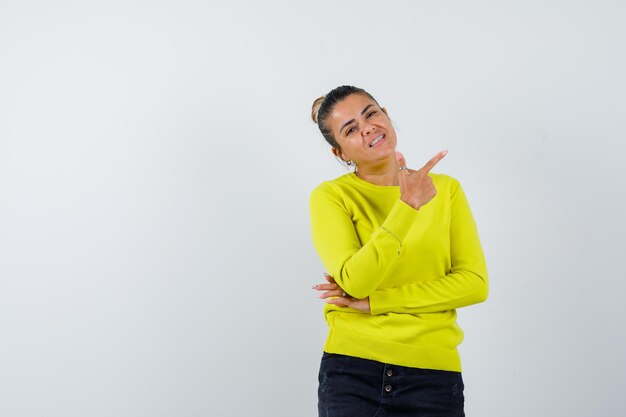 Jeune femme pointant vers la droite avec l'index en pull jaune et pantalon noir et l'air heureux