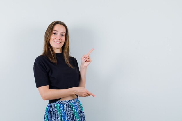 Jeune femme pointant vers la droite et vers le haut avec l'index en t-shirt noir et jupe bleue et à heureux