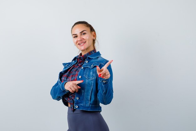 Jeune femme pointant vers l'extérieur en chemise à carreaux, veste, pantalon et à la gaieté. vue de face.