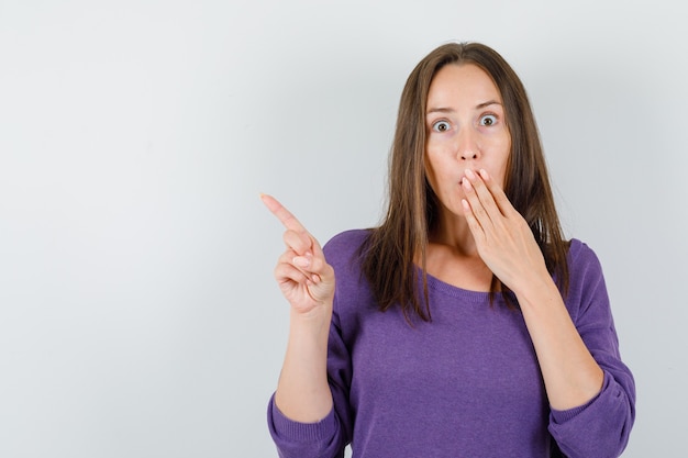 Jeune femme pointant vers l'extérieur tout en regardant en chemise violette et à la surprise. vue de face.