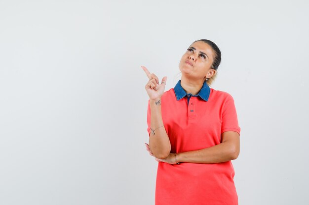 Jeune femme pointant vers la gauche avec l'index en chemise rouge et à la pensif. vue de face.