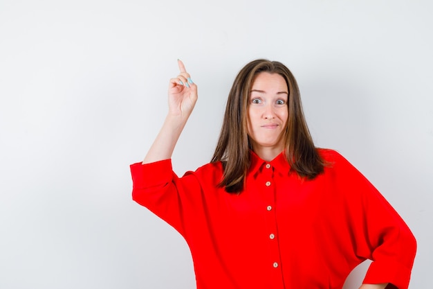 Photo gratuite jeune femme pointant vers le haut en blouse rouge et à la curieuse , vue de face.