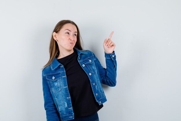 Jeune femme pointant vers le haut en blouse, veste et hésitante. vue de face.