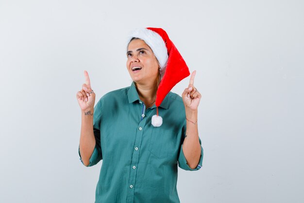 Jeune femme pointant vers le haut en chemise, bonnet de Noel et regardant joyeux, vue de face.