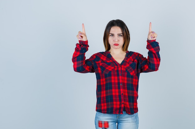 Photo gratuite jeune femme pointant vers le haut en chemise à carreaux, jeans et semblant s'ennuyer. vue de face.