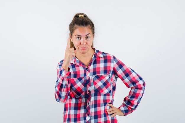 Jeune femme pointant vers le haut en chemise à carreaux et regardant confiant, vue de face.