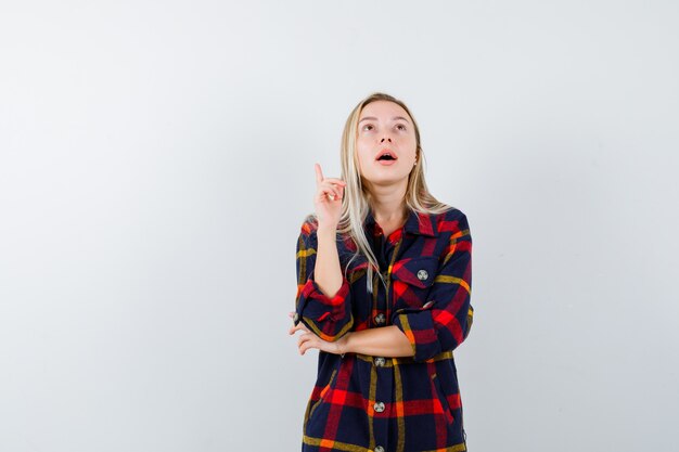 Jeune femme pointant vers le haut en chemise à carreaux et regardant perplexe, vue de face.