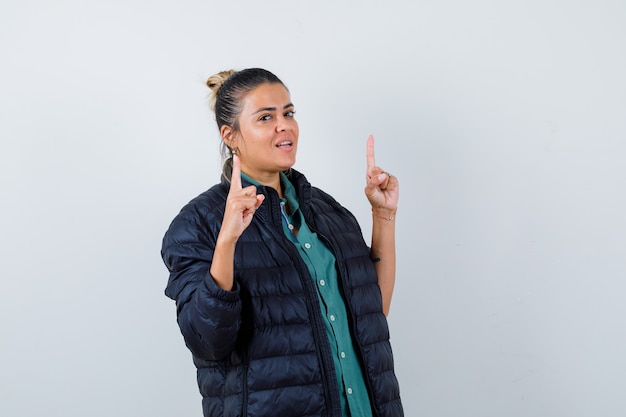 Jeune femme pointant vers le haut en chemise, doudoune et à l'air confiant, vue de face.