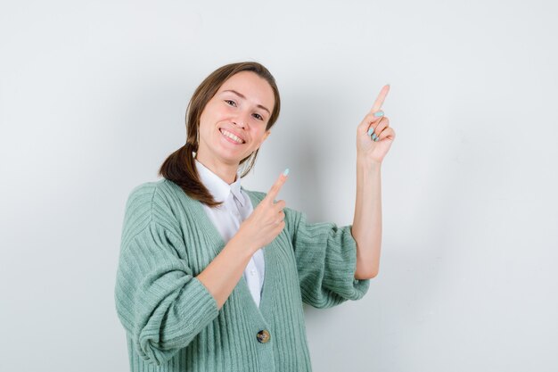 Jeune femme pointant vers le haut en chemisier, cardigan et l'air heureux, vue de face.