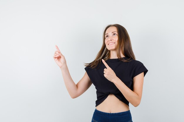 Jeune femme pointant vers le haut en chemisier noir, pantalon et à la joyeuse.