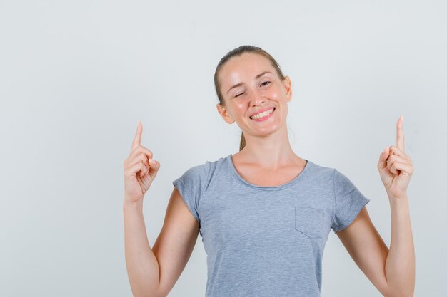 Jeune femme pointant vers le haut et clignant des yeux en t-shirt gris, vue de face.